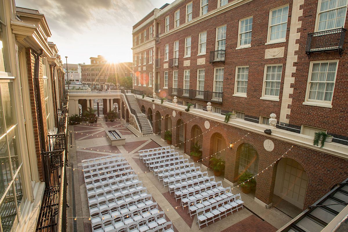 The image shows a courtyard set up for an event with rows of white chairs, surrounded by brick buildings, and illuminated by hanging lights.