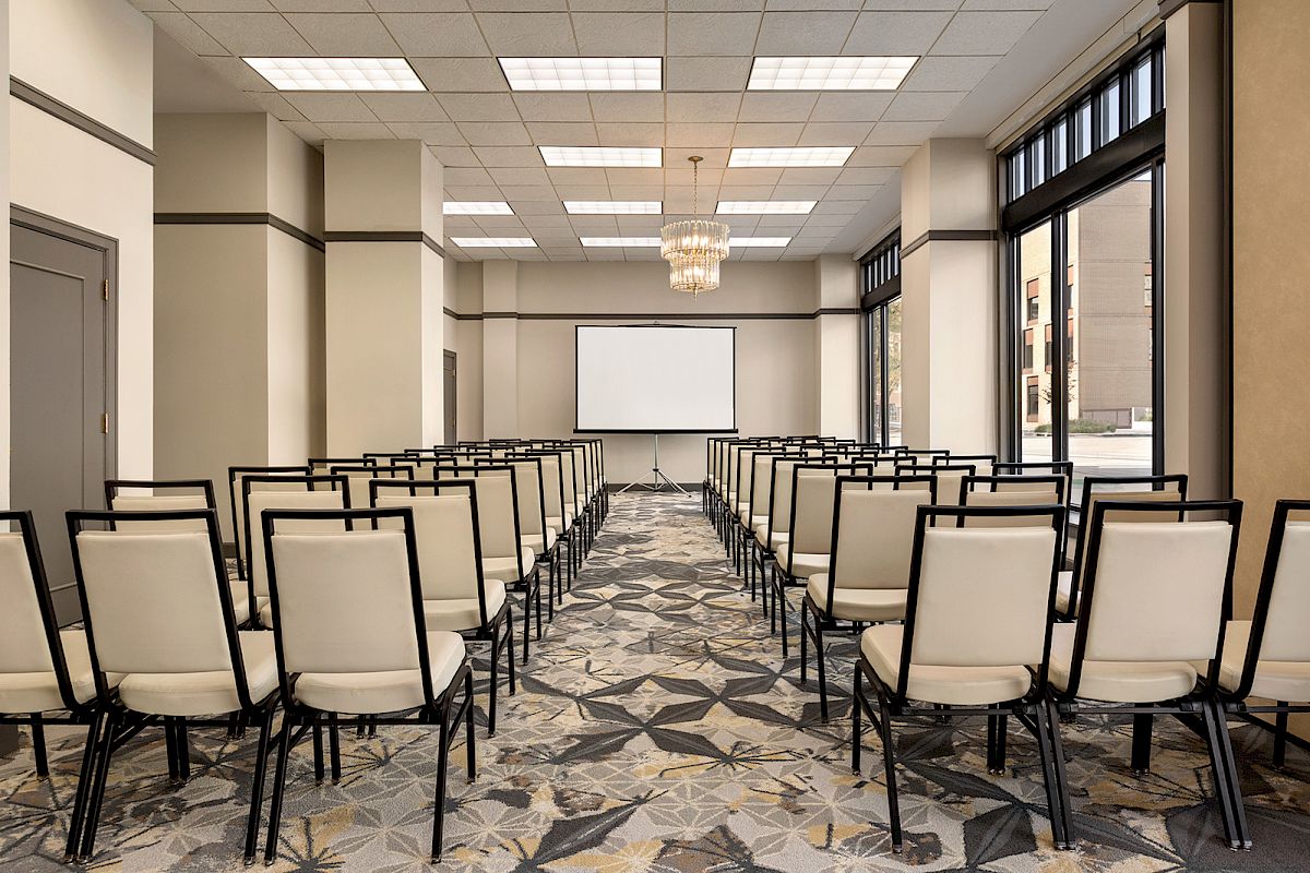 The image shows a conference room with rows of beige chairs facing a screen at the front, with large windows and a patterned carpet.