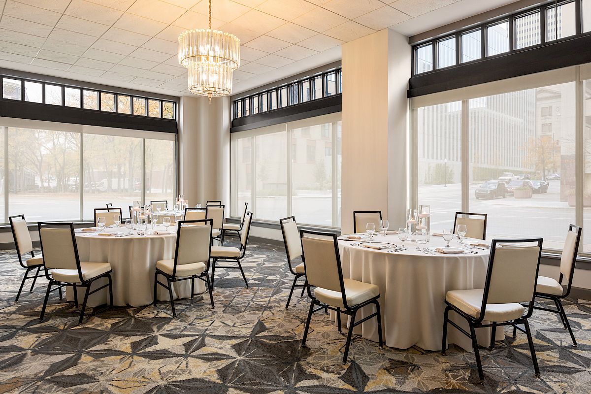 The image shows a banquet room with round tables set for an event, featuring chairs, place settings, and a chandelier, all in a bright space.