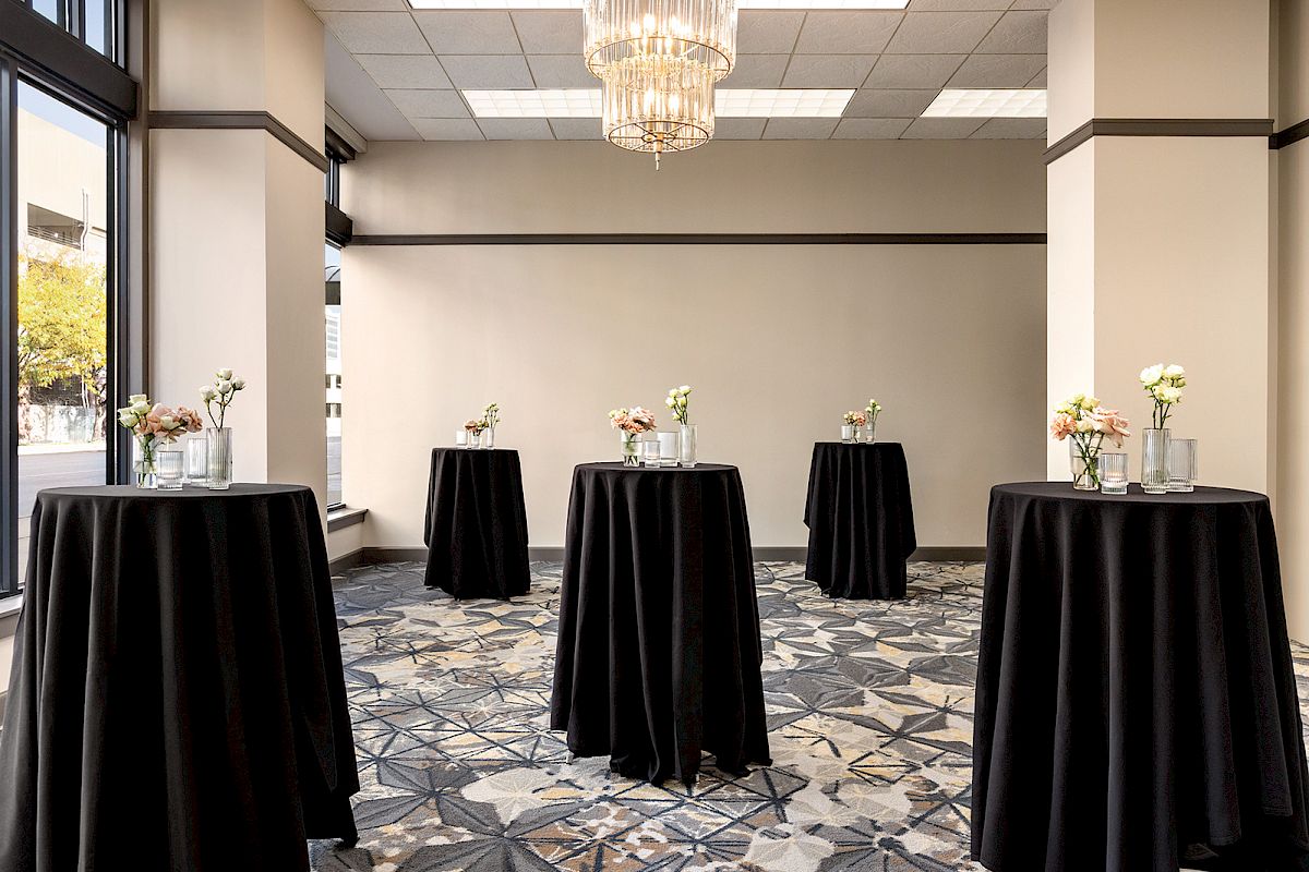 The image shows an elegant room setup with tall tables covered in black cloth, each adorned with small floral arrangements.