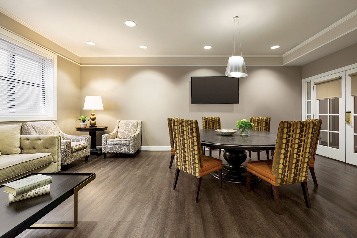 The image shows a modern living room with a round dining table, chairs, a sofa, armchairs, a lamp, and a wall-mounted TV.