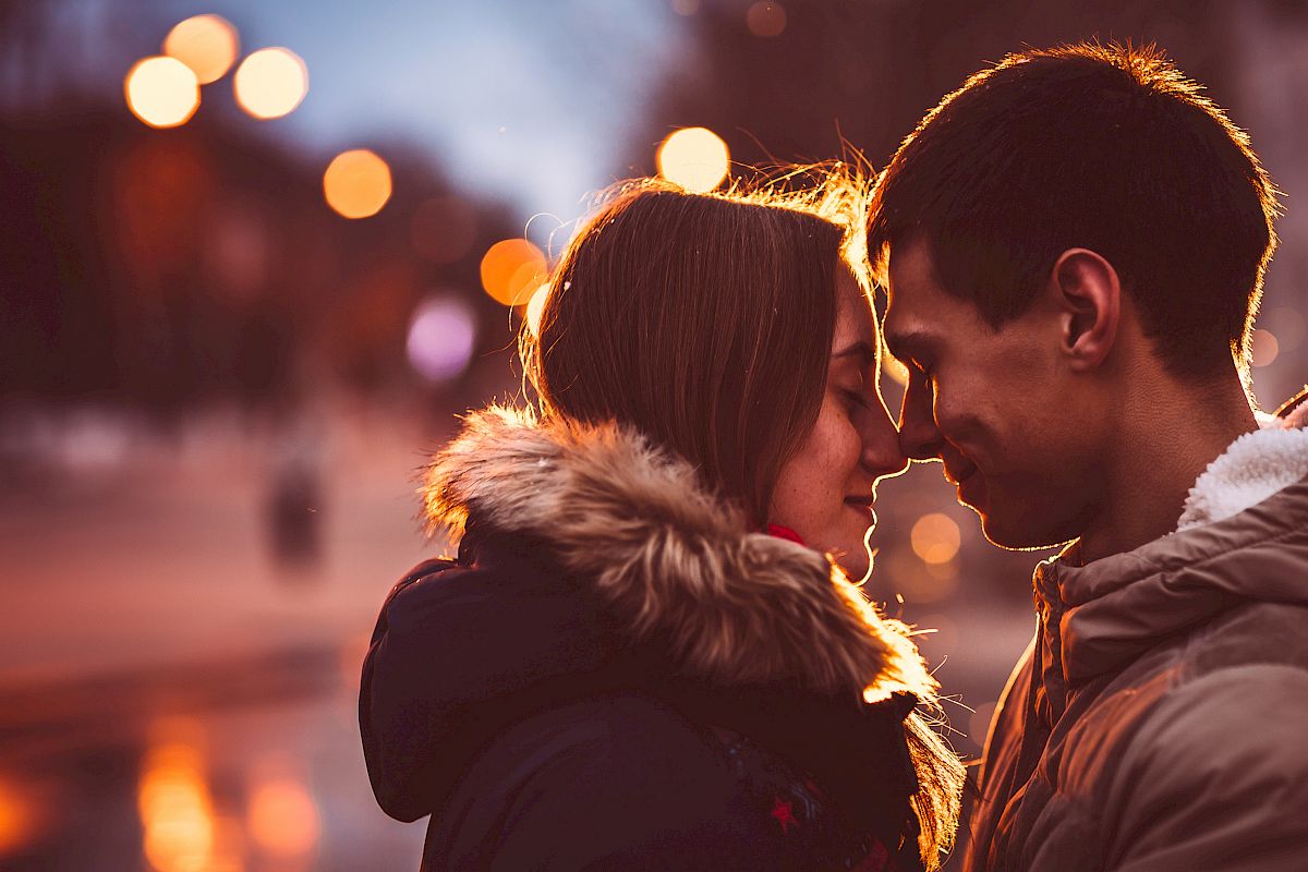 A couple in winter clothes stand closely together, gently touching foreheads, with a blurred, warmly lit urban background.