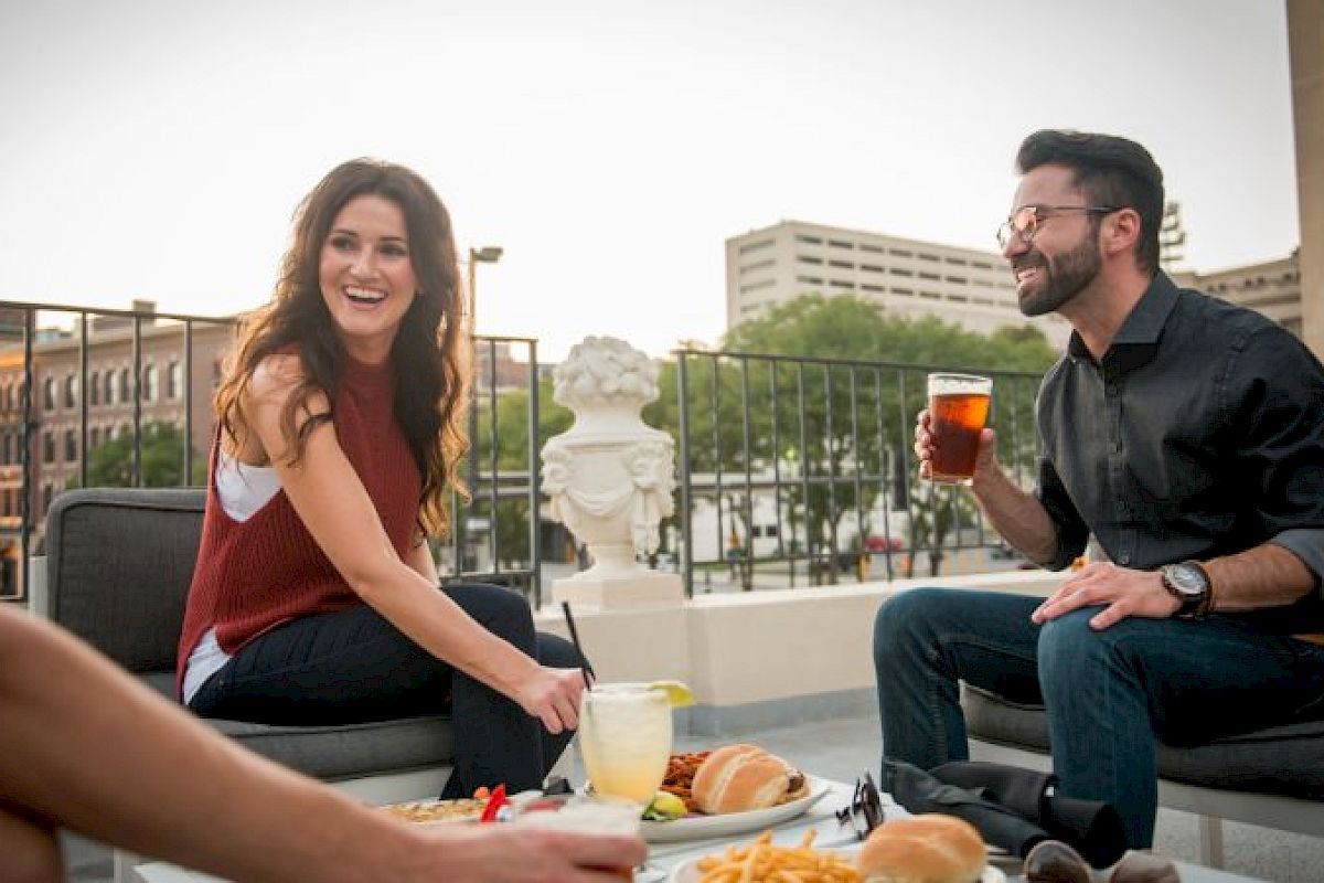 A group of people enjoying drinks and food on a rooftop patio, with city buildings in the background.