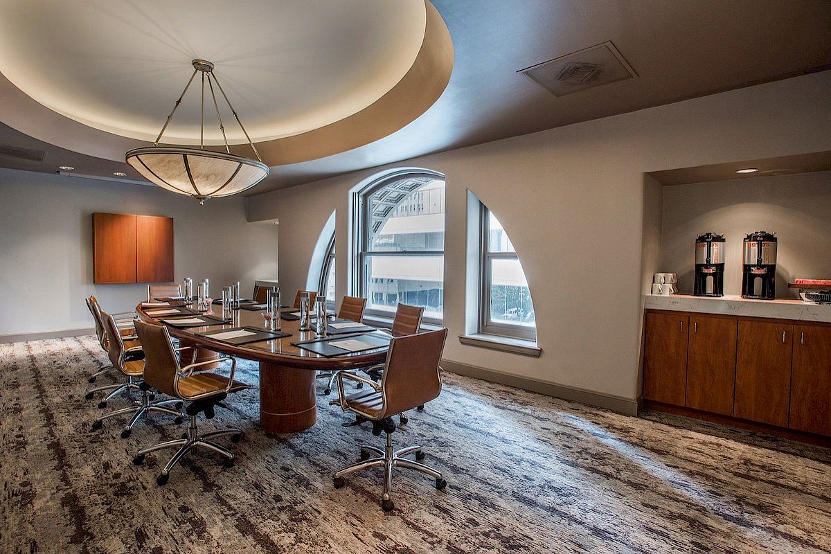 A modern conference room with a long table, office chairs, and a side counter with beverages. It has large windows and a stylish ceiling light.
