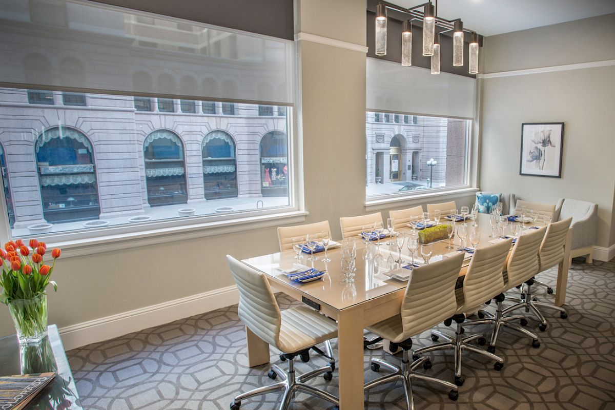 A modern conference room with a long table, chairs, glasses, and napkins, featuring large windows and a city view.