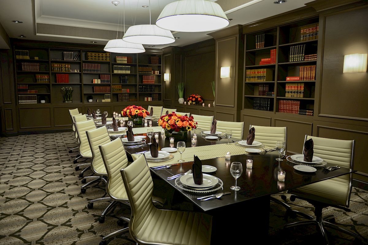 A dining room with a long table, elegant place settings, floral centerpieces, bookshelves, and pendant lighting.