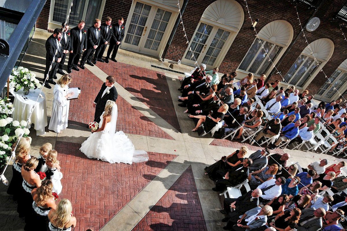 A wedding ceremony is taking place outdoors with a bride, groom, and officiant. Guests are seated, and the wedding party stands nearby.