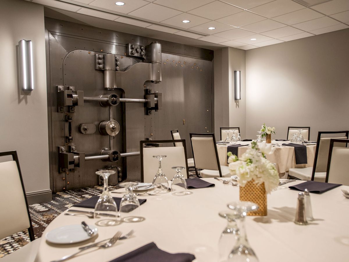 The image shows a dining room with tables set for a meal in front of a large, ornate vault door.