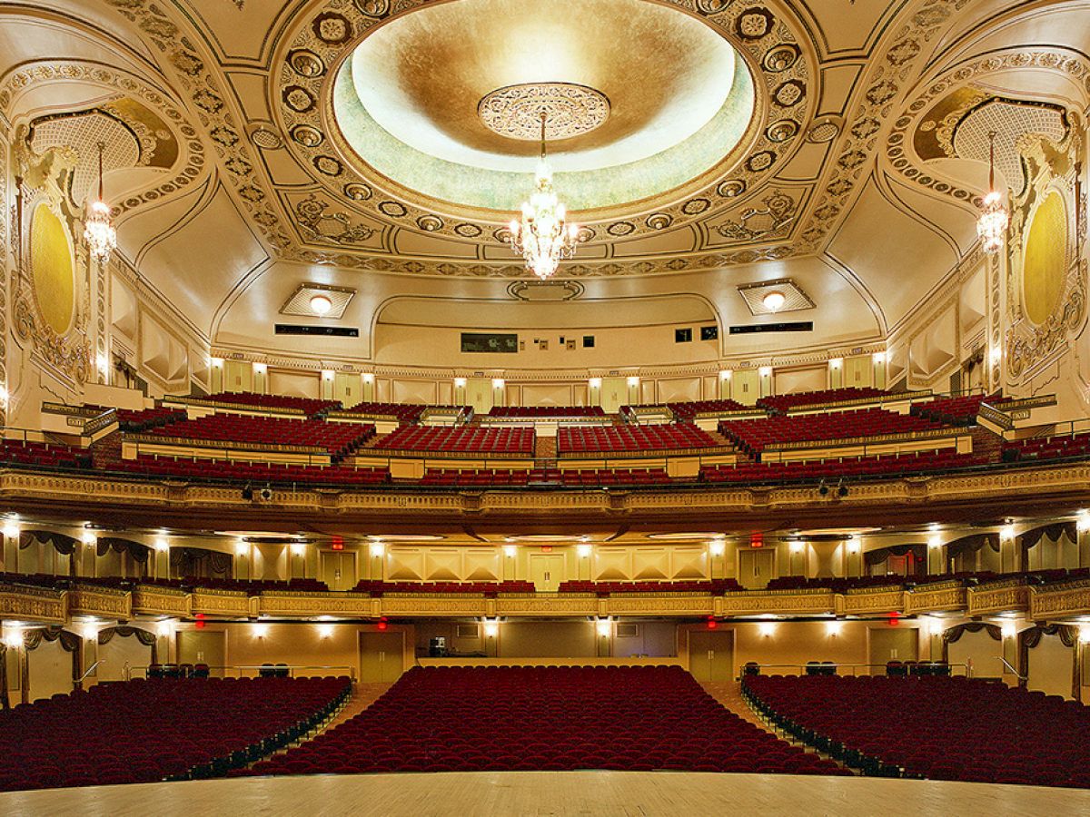 The image shows an ornate, empty theater with multiple seating levels, elegant chandeliers, and decorative details on the ceiling and walls.