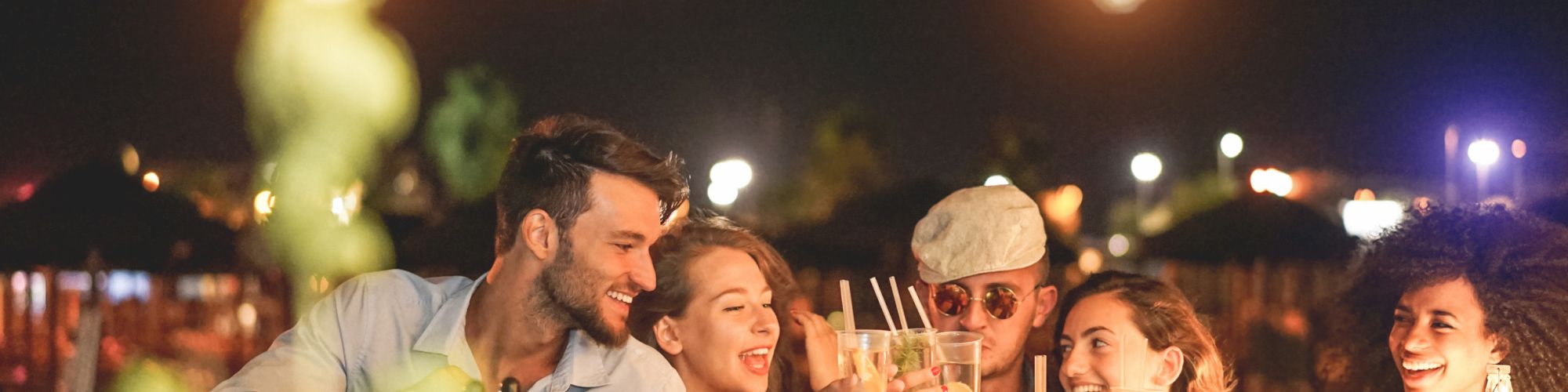 A group of friends enjoying drinks and smiling together at a bar, with festive string lights in the foreground and a lively night scene behind.