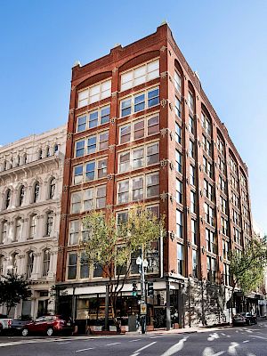 A corner view of a red brick multi-story building with large windows and trees at the base, located at a city intersection.