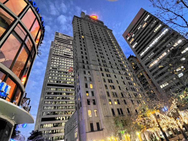 The image shows a low-angle view of tall buildings in a city at dusk, with lights illuminated and trees partially visible, creating an urban scene.