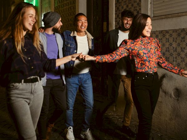 A group of five people are happily walking down an outdoor street at night, with two of them holding hands and smiling.