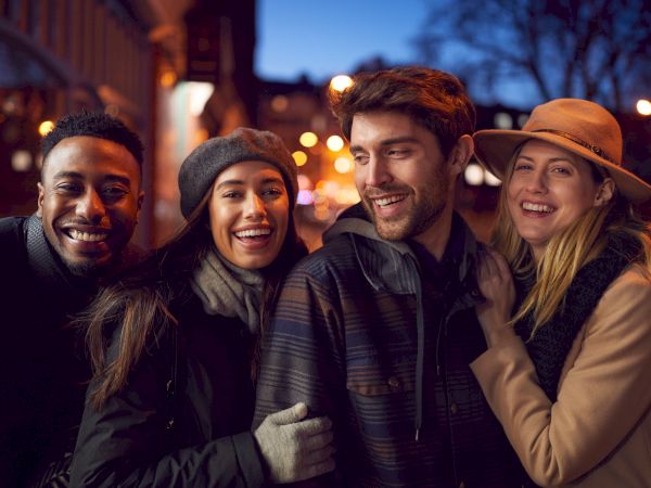 A group of four people, bundled in winter clothing, smiling and enjoying an evening out on a street with blurred lights in the background.