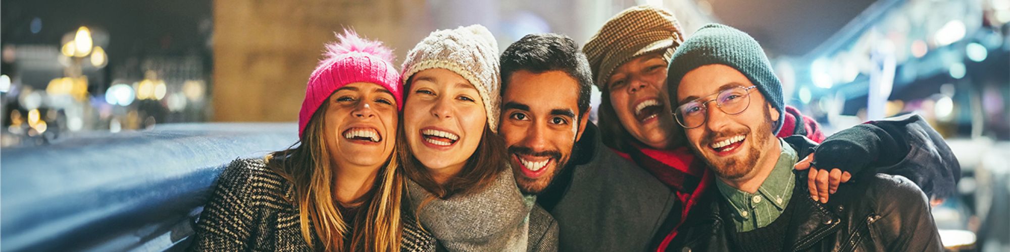A group of five friends smiling and wearing winter clothes, standing outdoors at night with a bridge and city lights in the background.