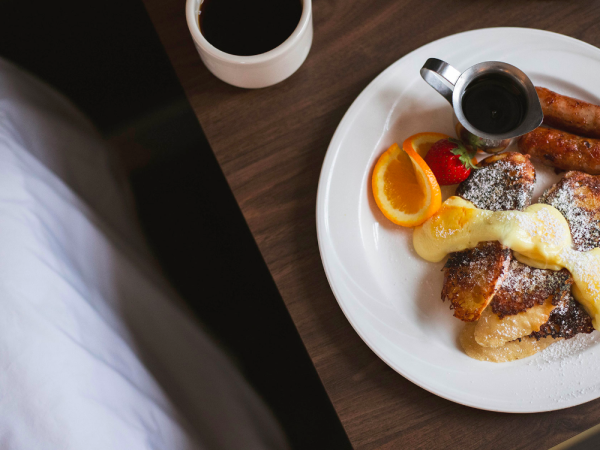 A plate with French toast, sausages, orange slices, a strawberry, and syrup is on a table next to a cup of coffee.