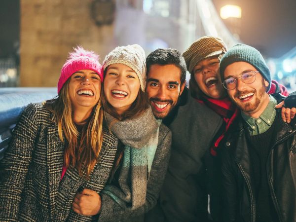 A group of five friends is smiling and posing for a photo on a bridge at night, dressed warmly in winter clothing and hats.