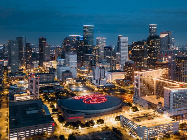 A city skyline at dusk with illuminated buildings and a round building in the foreground featuring a bright red logo on its roof.