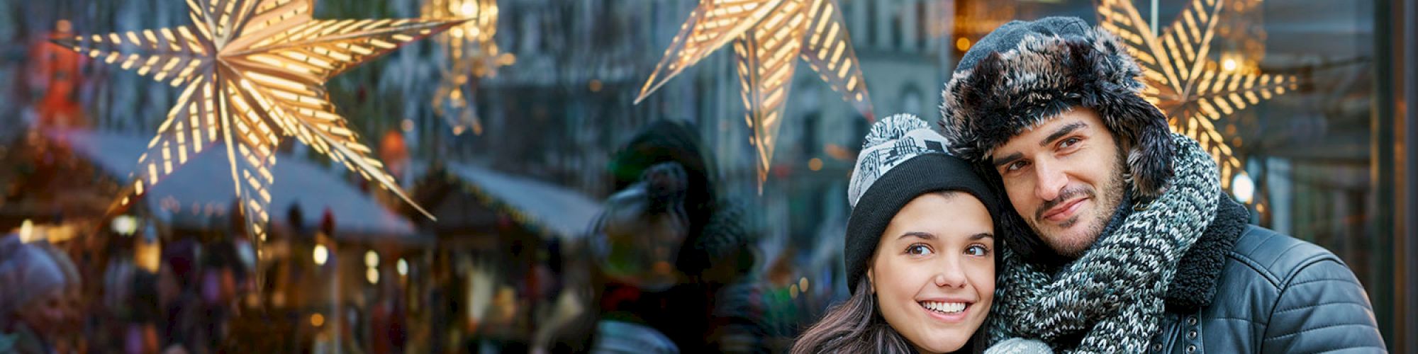 A couple is posing with a shopping bag in front of a festive window display featuring decorative stars and lights.