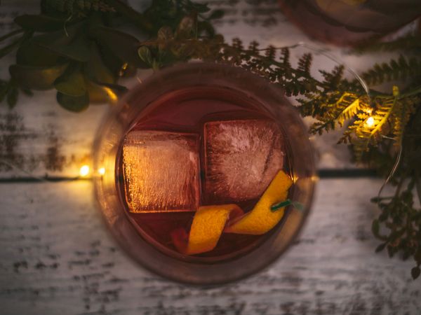 A beverage in a glass with ice cubes and a lemon twist, surrounded by greenery and string lights on a wooden surface.