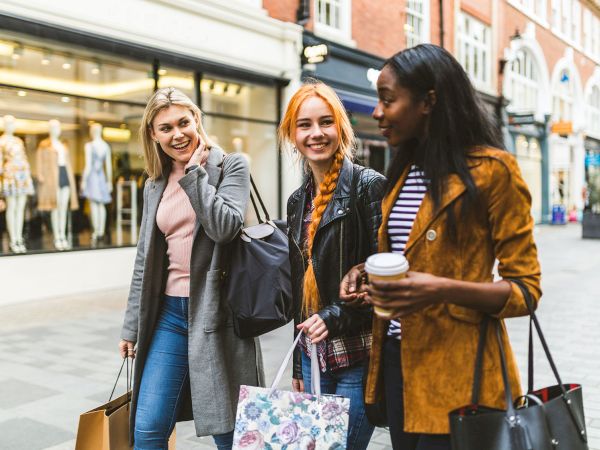 Three women are walking with shopping bags, smiling and conversing, in an urban setting with storefronts in the background.