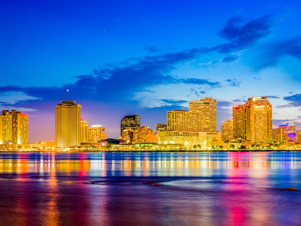 A vibrant cityscape at dusk with illuminated buildings reflecting on the water. The sky transitions from blue to warm shades.