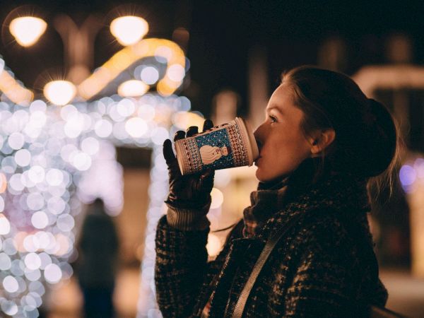 A person drinks from a festive cup against a backdrop of blurred holiday lights in the evening.