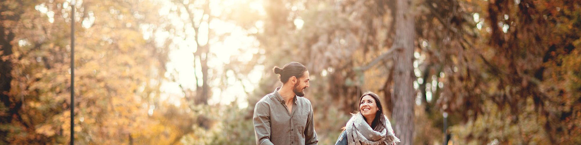 A couple is walking and smiling in a park during autumn, surrounded by fallen leaves and trees.