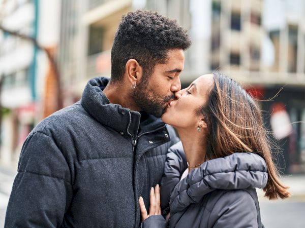 A couple kissing outdoors, both wearing winter jackets, with a city street blurred in the background.
