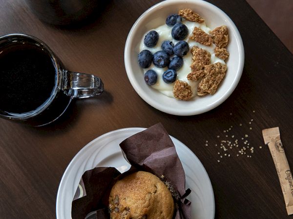 A cup of coffee, a bowl of yogurt with blueberries and granola, a muffin on a plate, sugar packet, and some spilled sugar on a wooden table.