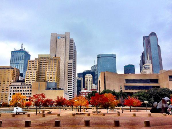 A skyline with tall buildings in the background and a plaza with trees displaying fall colors in the foreground under a cloudy sky.