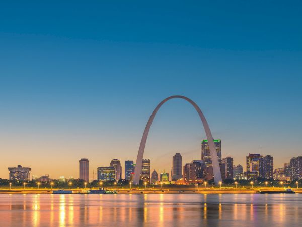 The image shows the skyline of a city at dusk featuring a large, distinctive arch near the waterfront with lights reflecting in the water.