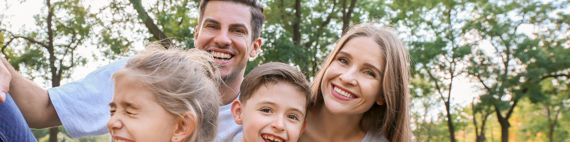 A cheerful family of four sits together on grass in a park, smiling and enjoying each other's company on a sunny day.