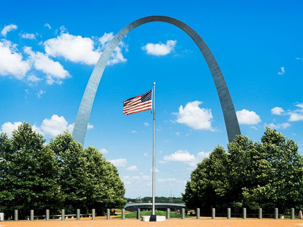The image shows the Gateway Arch in St. Louis with an American flag in front, set against a blue sky with scattered clouds.
