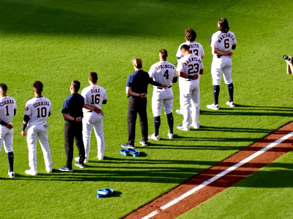 Baseball players line up on the field, with their hats off. A photographer crouches nearby, capturing the moment.