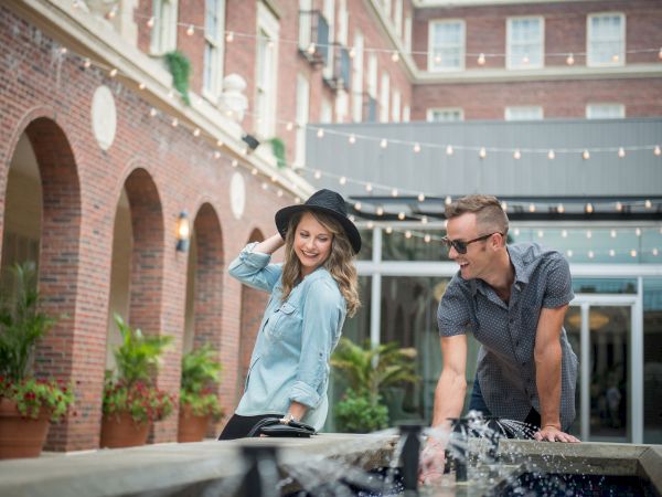 A man and woman are enjoying themselves near a fountain in a courtyard with string lights and a brick building in the background.