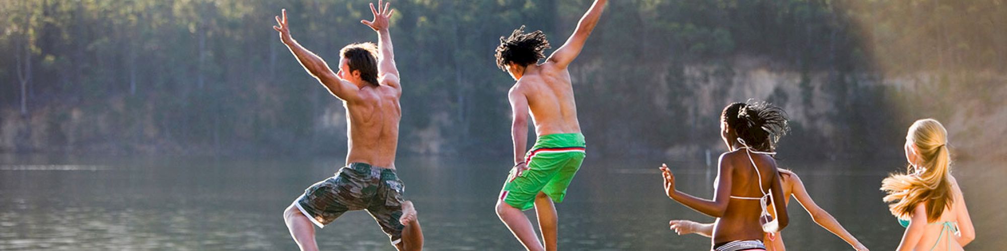 A group of young people are joyfully jumping off a wooden dock into a lake, surrounded by trees and calm water.