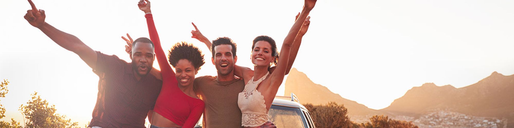 Four people happily posing next to a car on a scenic road, raising their arms in celebration as the sun sets in the background.