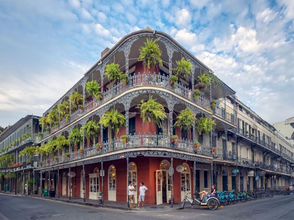 A historic building with ornate balconies and lush greenery on a street corner, with people and a bicycle rickshaw nearby.