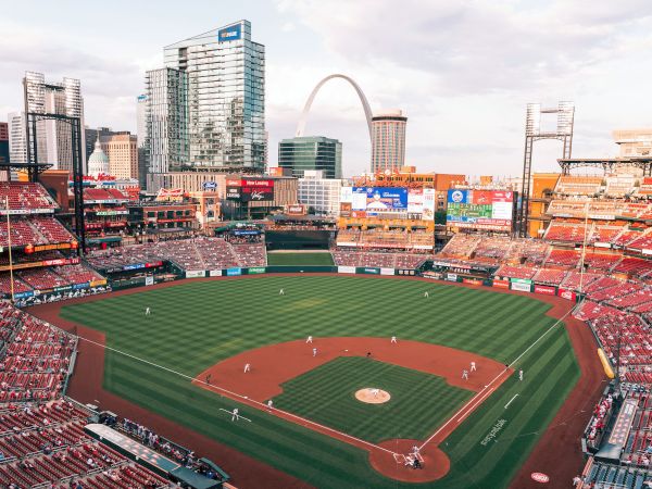 A baseball stadium filled with fans, with a game in progress. The city skyline and a large arch are visible in the background.