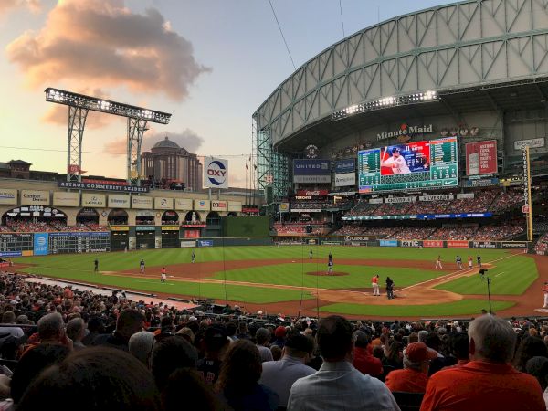 A baseball game is in progress at a stadium with a closed roof. Spectators fill the seats, and the field is lit by the setting sun.