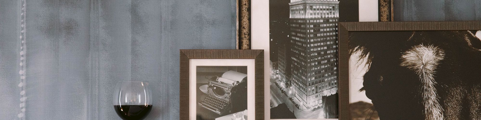 A glass of red wine and three framed black-and-white photos on a shelf against a textured wall.