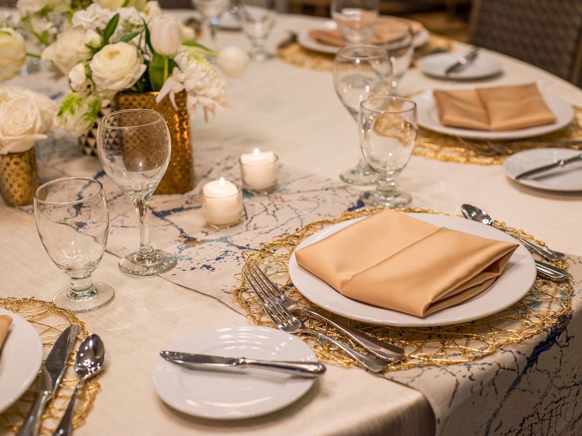 Elegant table setting with beige napkins, wine glasses, white flowers, and candles on a decorative tablecloth.