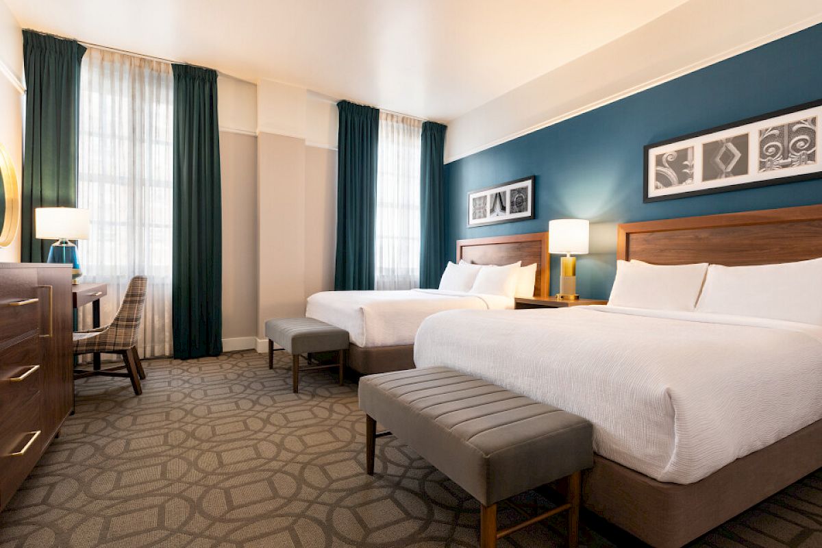 The image shows a hotel room with two beds, a desk, table lamp, chair, decorative art on the wall, and large windows with curtains.