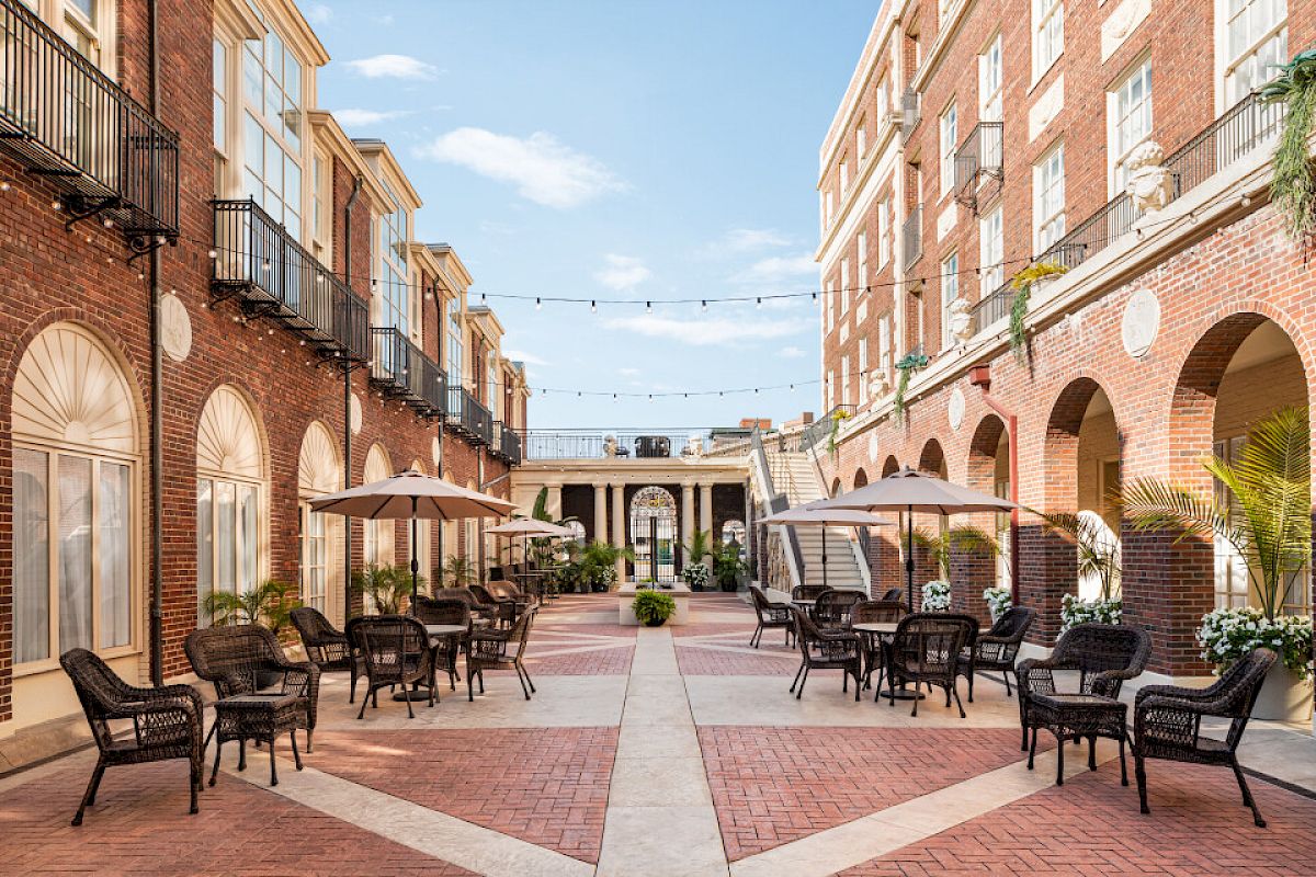 An outdoor courtyard with brick buildings, archways, tables, chairs, and umbrellas. String lights hang across the sky.