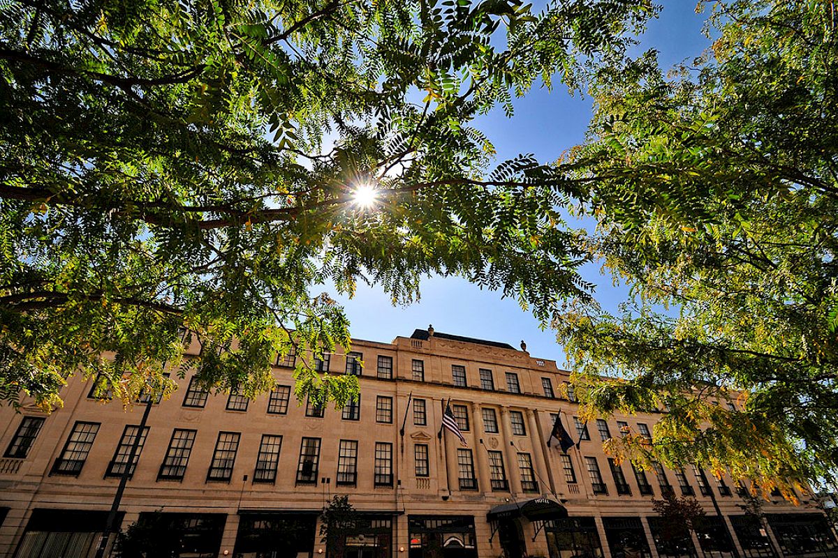A grand building is seen through leafy trees with sunlight streaming through, against a clear blue sky.