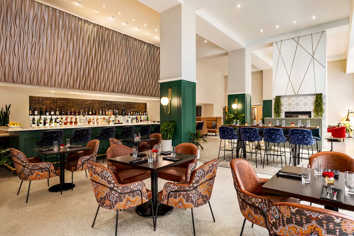 The image shows a modern, stylish restaurant interior with patterned chairs, a well-stocked bar, and elegant lighting fixtures.