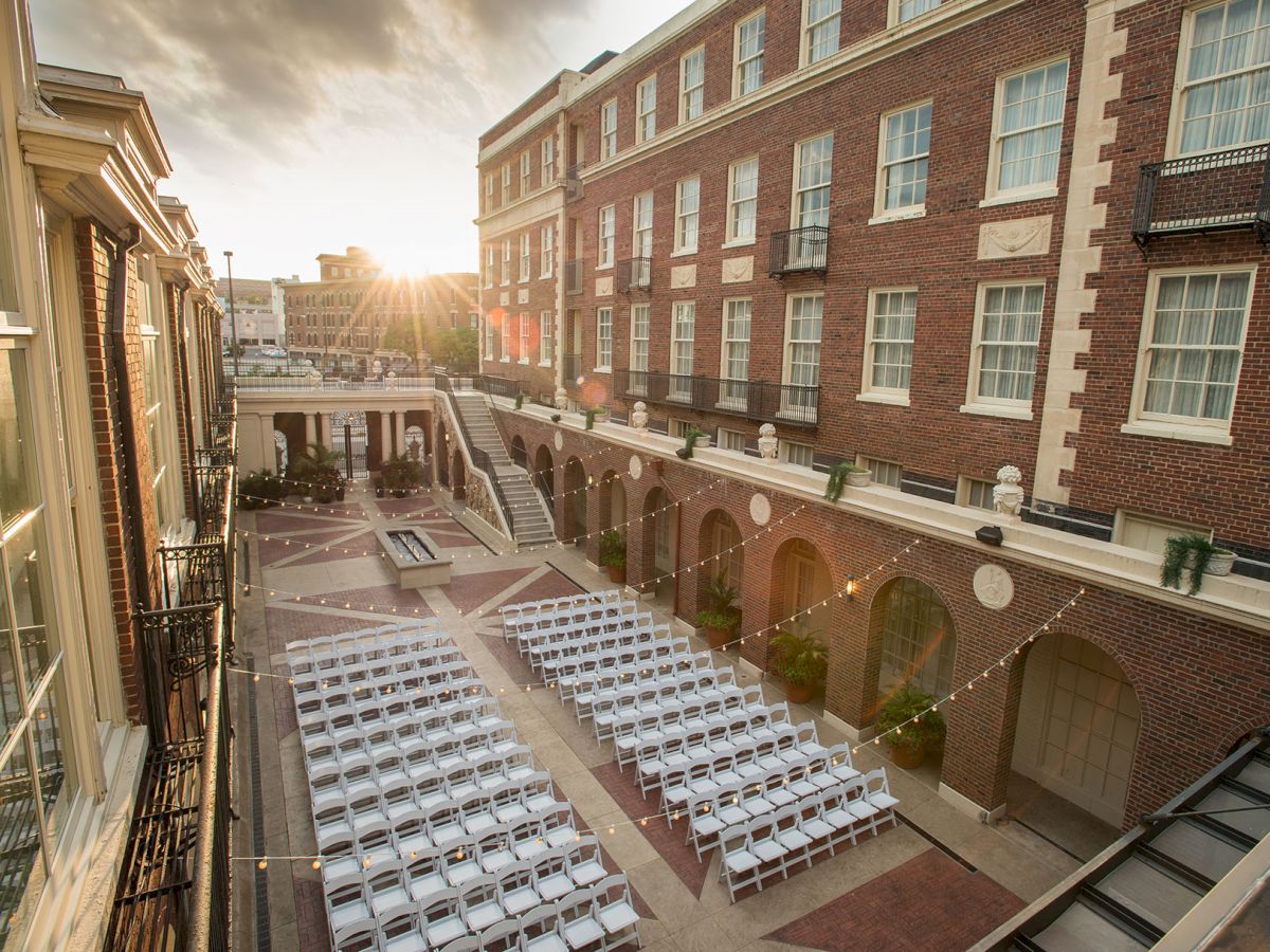 An outdoor venue with white chairs set up for an event, surrounded by a brick building, with string lights overhead and the sun setting.