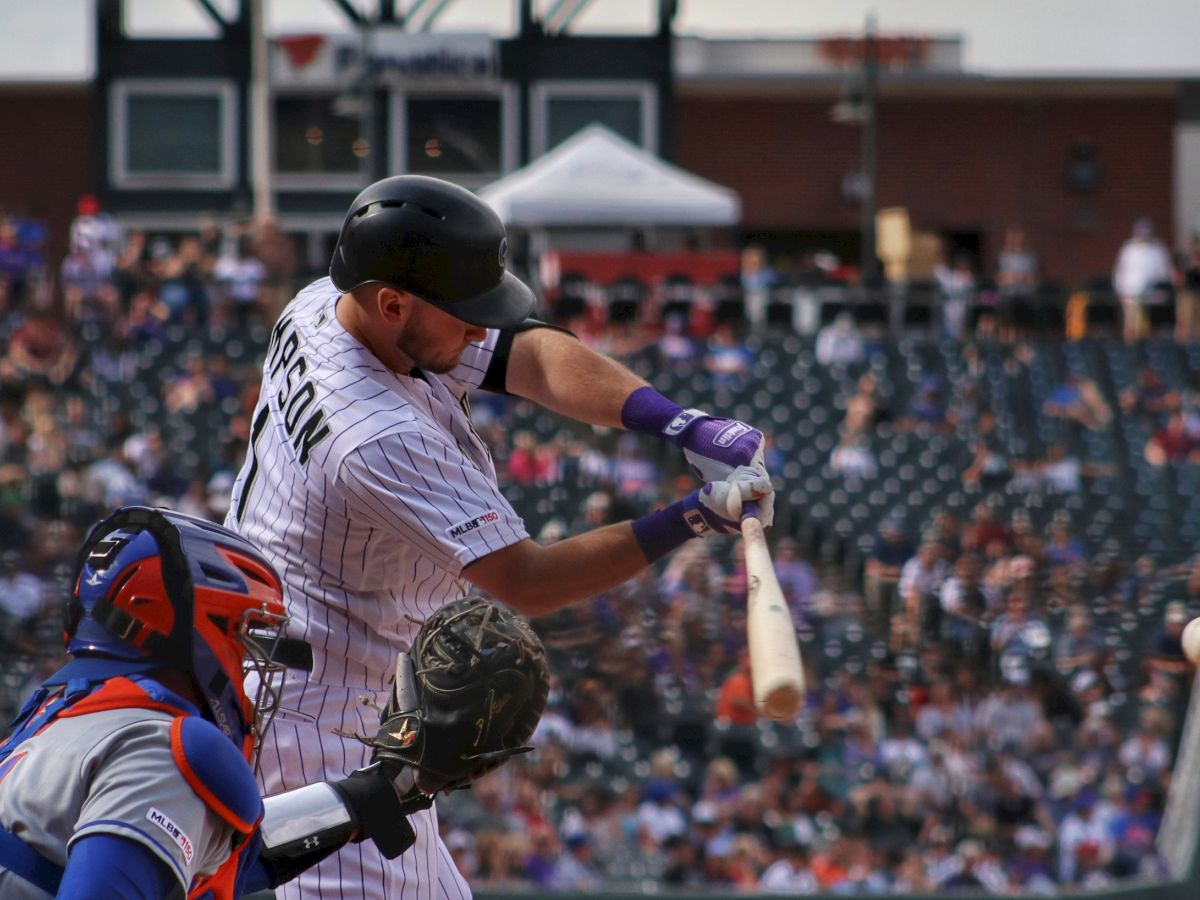 A baseball player in a white jersey swings at a pitch while a catcher, in blue gear, prepares to catch the ball in a crowded stadium.