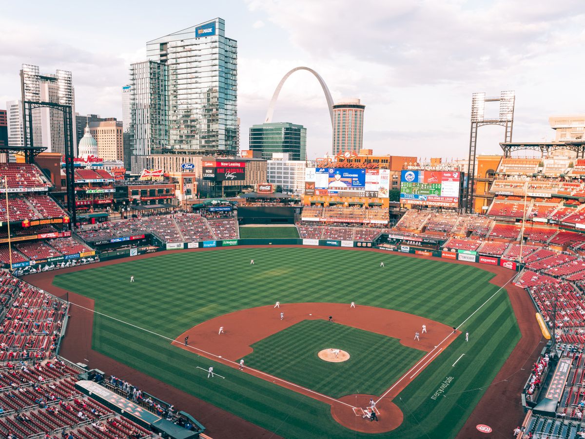 A baseball stadium with a game in progress, surrounded by city buildings and the iconic Gateway Arch in the background.